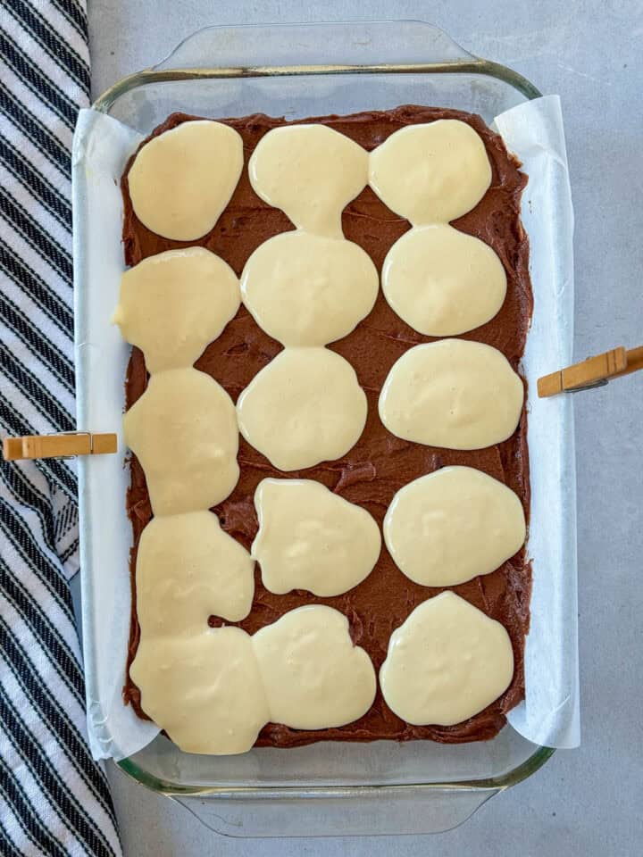 Cheesecake batter dolloped on top of brownie layer in glass pan.