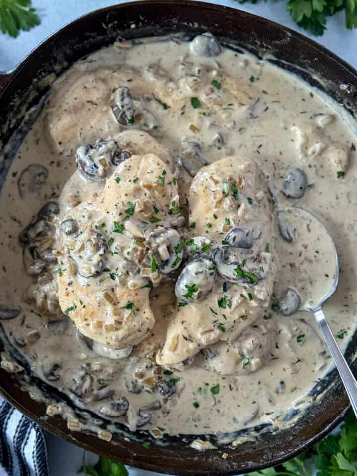 Top view of chicken breasts in creamy mushroom sauce with mushrooms atop of the chicken in a skillet and a spoon in the skillet.
