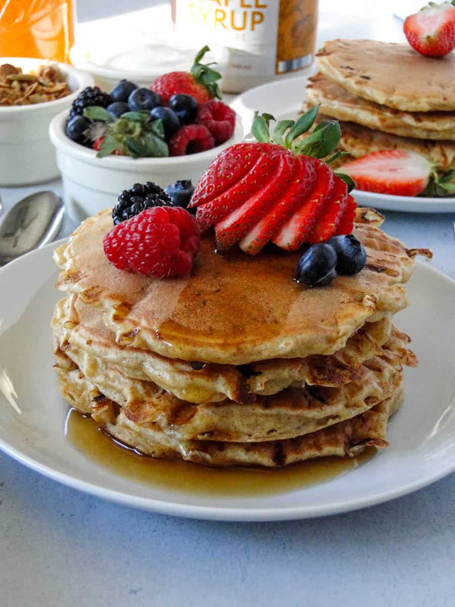 Close up side view of four cottage cheese pancakes stacked on white round plate topped with maple syrup and berries.
