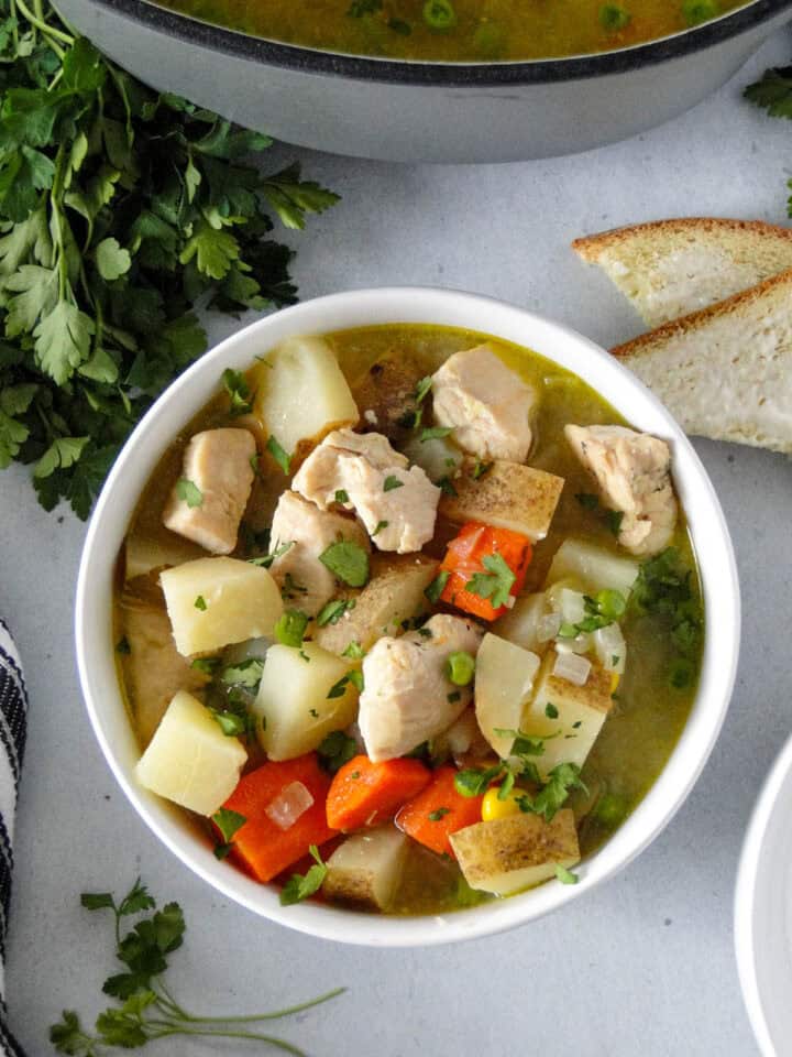 Chicken and potato stew in white round bowl in front of dutch oven with buttered bread triangles on the side.