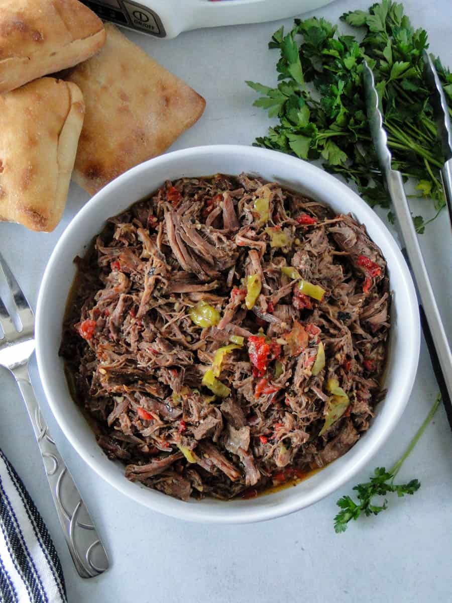 Shredded crock pot Italian beef in white serving bowl surrounded by ciabatta buns.