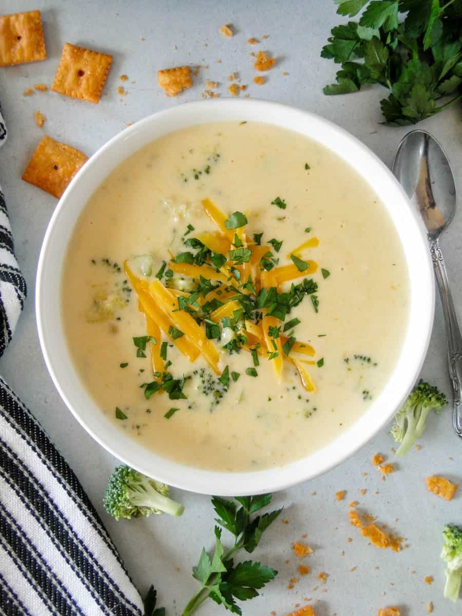 Top view of cheesy broccoli soup in white round bowl garnished with extra shredded cheese and parsley.