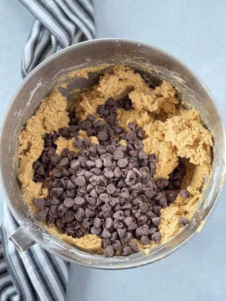 Chocolate chips added to mixed cookie dough in metal mixing bowl.
