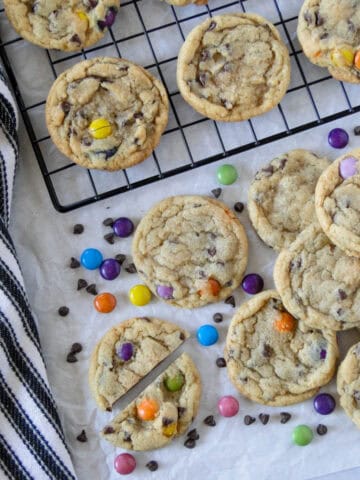 Chocolate chip m&m cookies on wire rack in in pile in front of rack with one cookie cut in half.