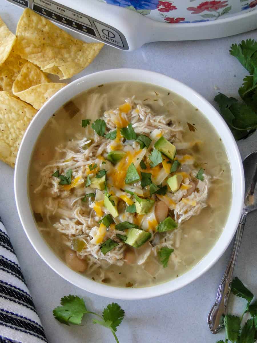 Top view of crock pot white chicken chili in white round bowl with tortilla chips and the crock pot on the side.