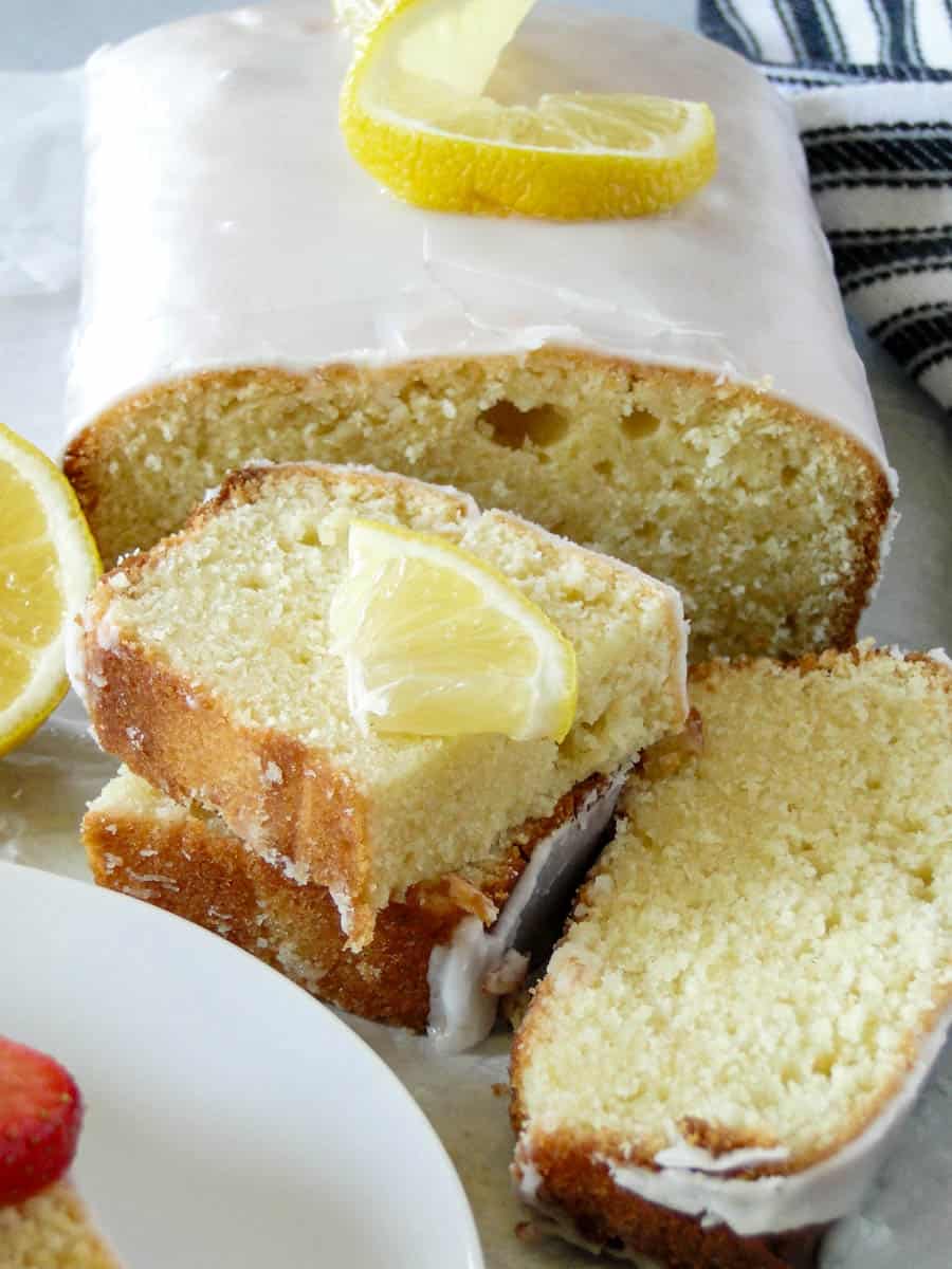 Front view of glazed lemon pound cake with slices stacked in front of loaf and topped with lemon triangle.