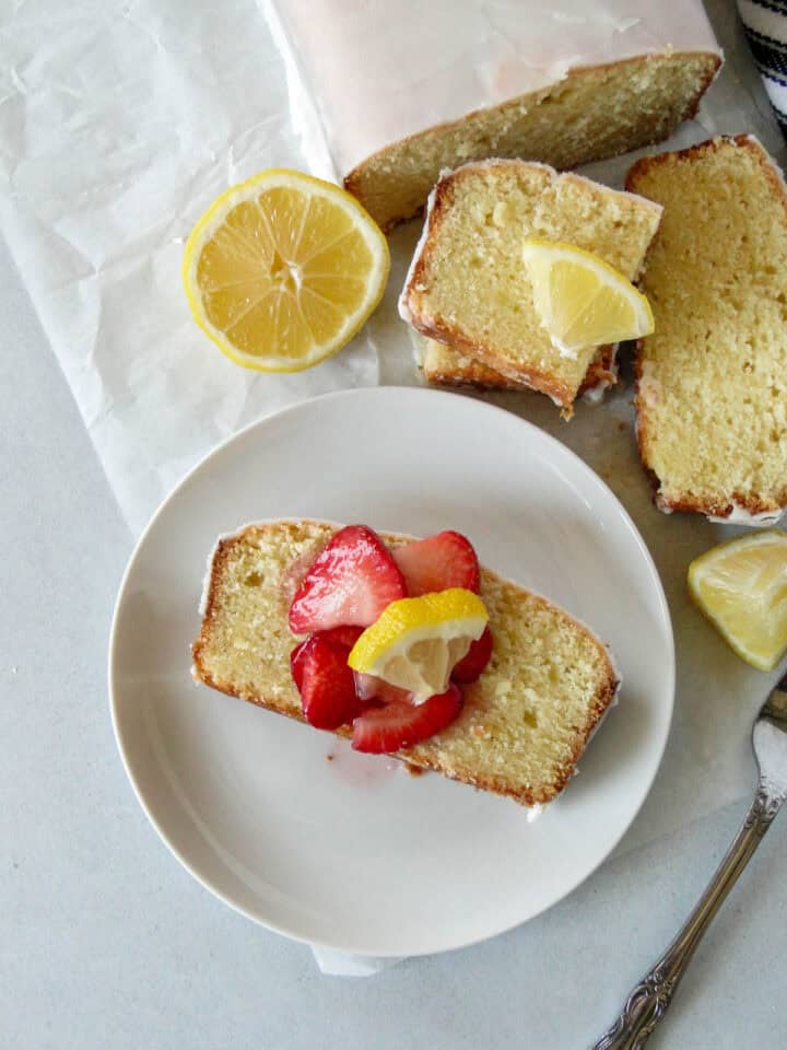 Slice of pound cake on white round plate topped with macerated strawberries and a small lemon triangle.