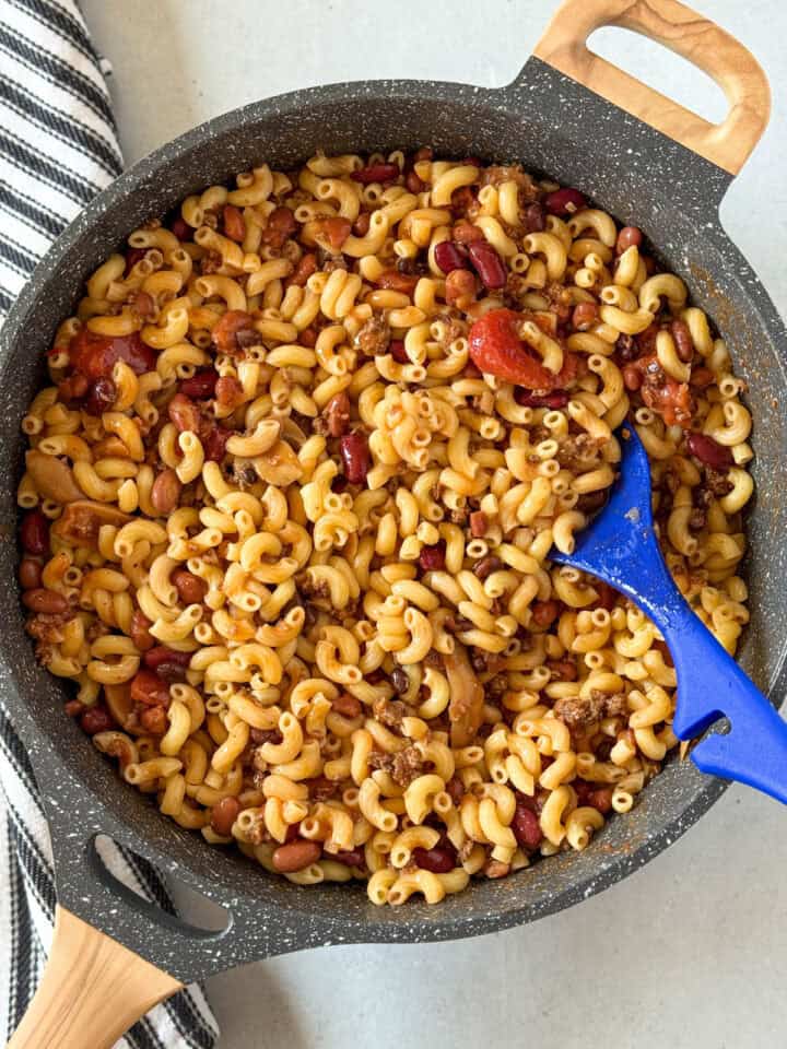 Leftover chili stirred into macaroni in skillet with blue spoon.