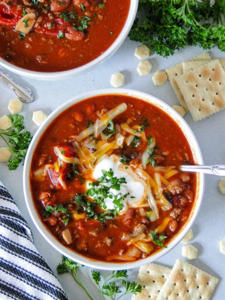 Top view of three bean chili in white round bowl with spoon and topped with cheese and sour cream.