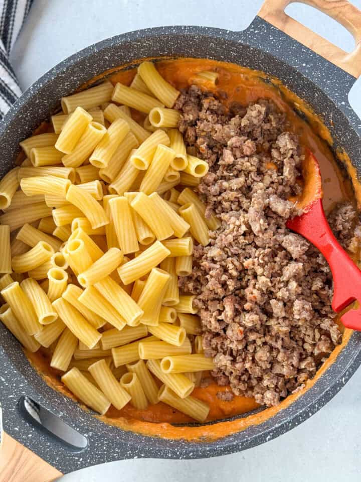 Cooked pasta and sausage added to sauce in large skillet with red spoon.