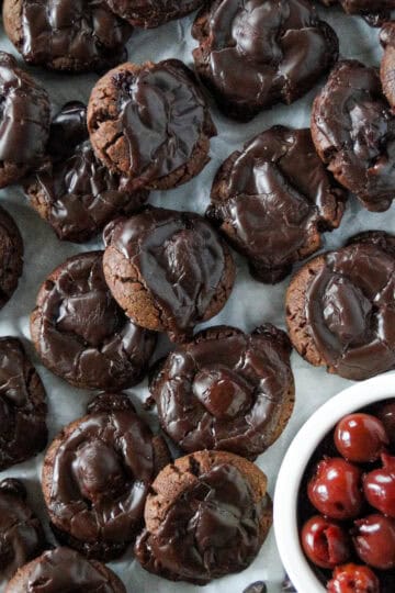 Top view of chocolate covered cherry cookies scattered and layered on parchment paper with a bowl of cherries in the bottom right corner.