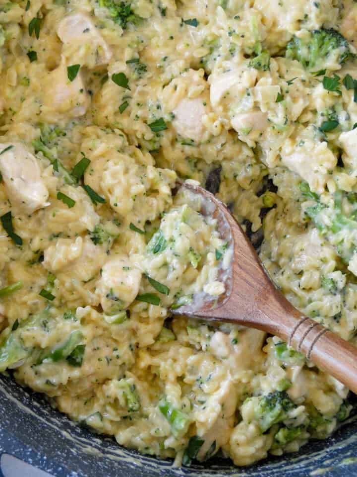 Close up top view of broccoli cheddar chicken orzo in skillet with wooden serving spoon.
