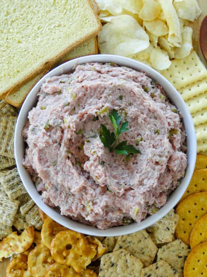 Top view of easy ham salad in white round bowl on board with crackers, chips and sliced of bread.