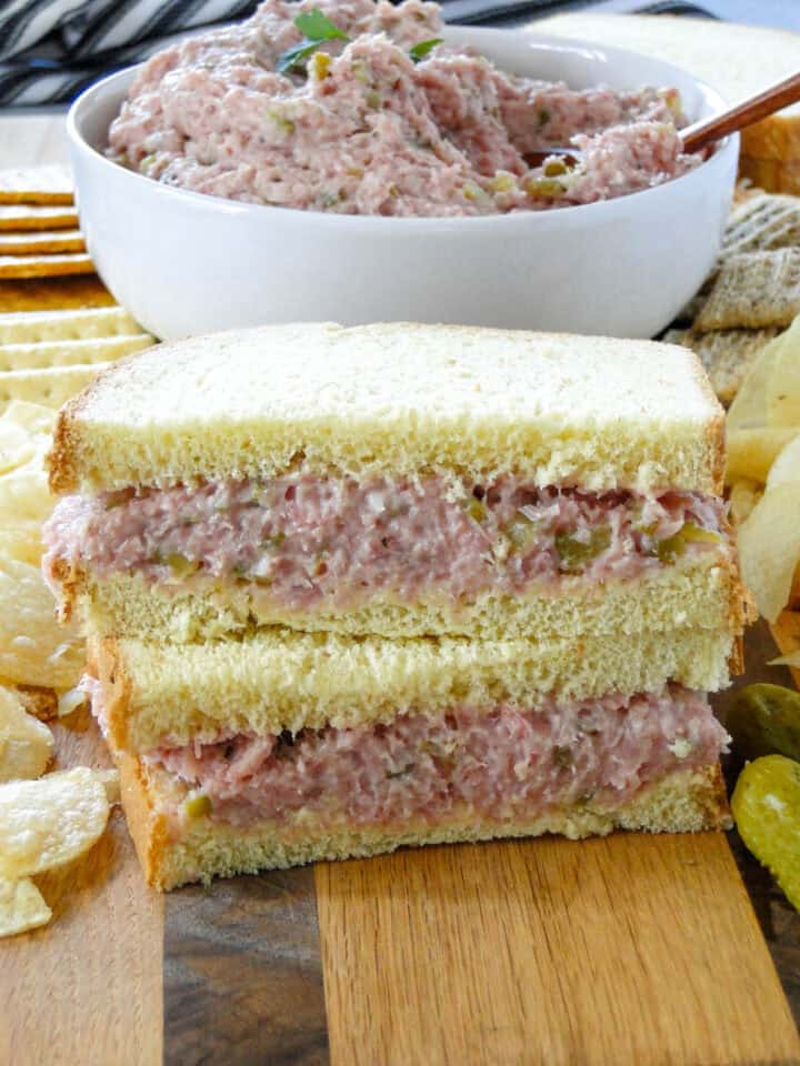 Close up side view of easy ham salad spread on bread to make a sandwich that is cut in half and stacked on board in front of bowl of ham salad.