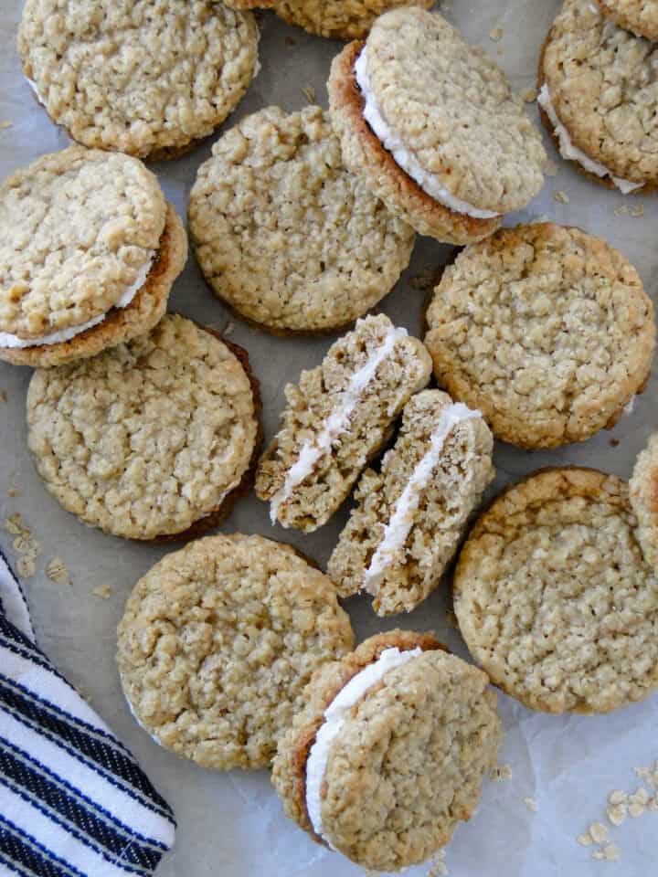 Homemade oatmeal cream pies scattered on parchment paper with one cookie cut in half and showing center of cookie.
