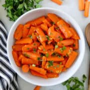 Honey glazed carrots in white round serving bowl with wooden serving spoon on the side.