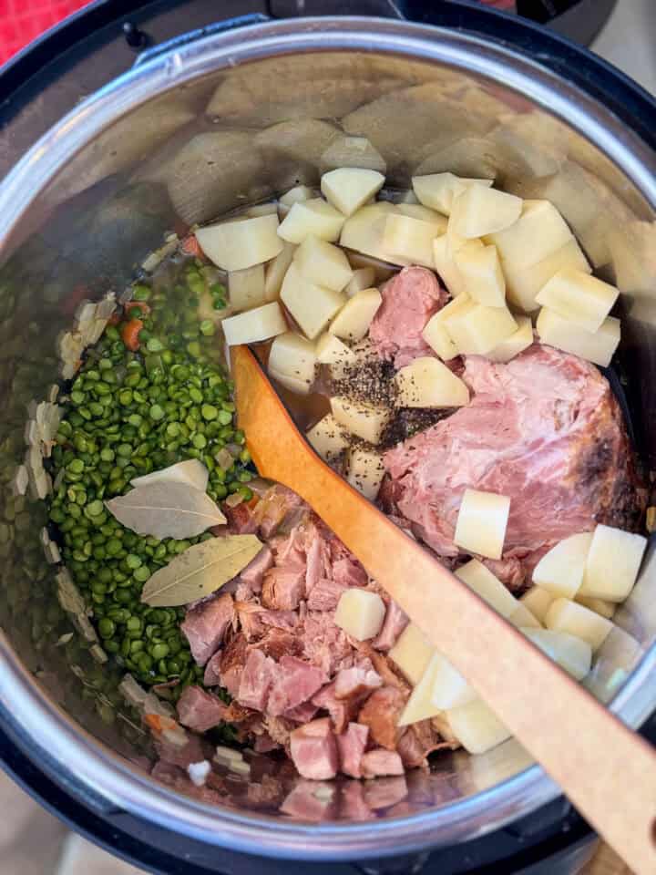 Split peas, ham, ham bone, potatoes, bay leaves and black pepper added to cooked veggies in instant pot with wooden spoon.