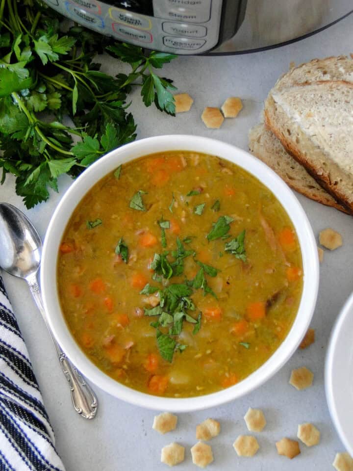 Top view of instant pot split pea soup in white round bowl with spoon and buttered bread to the side sitting in front of instant pot. 