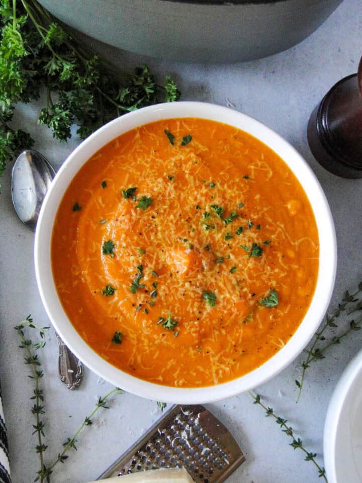 Creamy carrot soup in white round bowl garnished with parsley and parmesan cheese with spoon next to bowl.