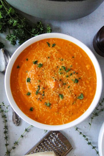 Creamy carrot soup in white round bowl garnished with parsley and parmesan cheese with spoon next to bowl.