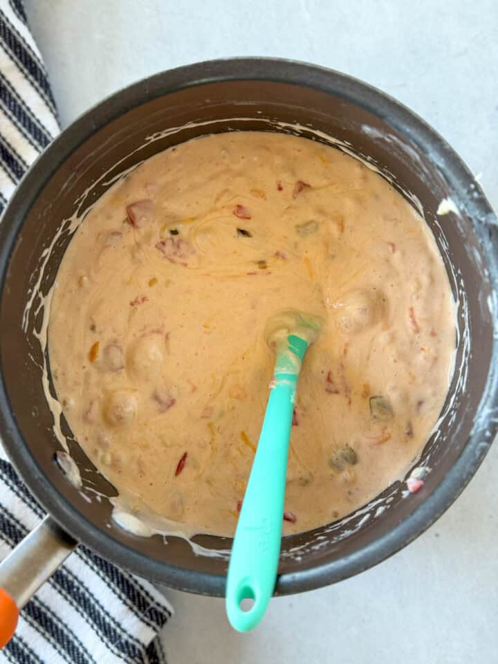 Heavy cream added to melted cheese in pot with green spatula.