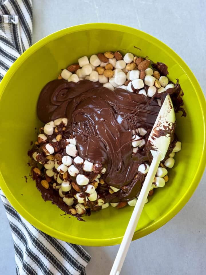 Chocolate poured over marshmallow and nuts in large green mixing bowl with white spatula.
