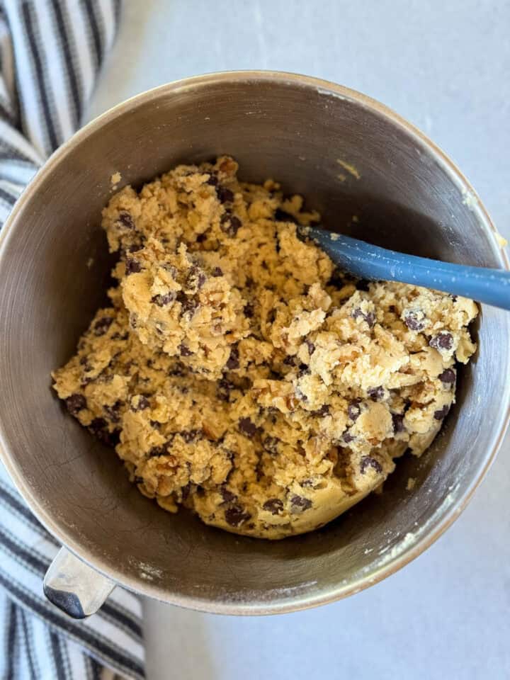 Walnuts and chocolate chips stirred into cookie dough with blue spatula in metal mixing bowl.