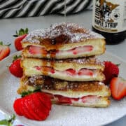 Side view of stuffed strawberry french toast cut in half, stacked and being drizzled with maple syrup on white round plate.
