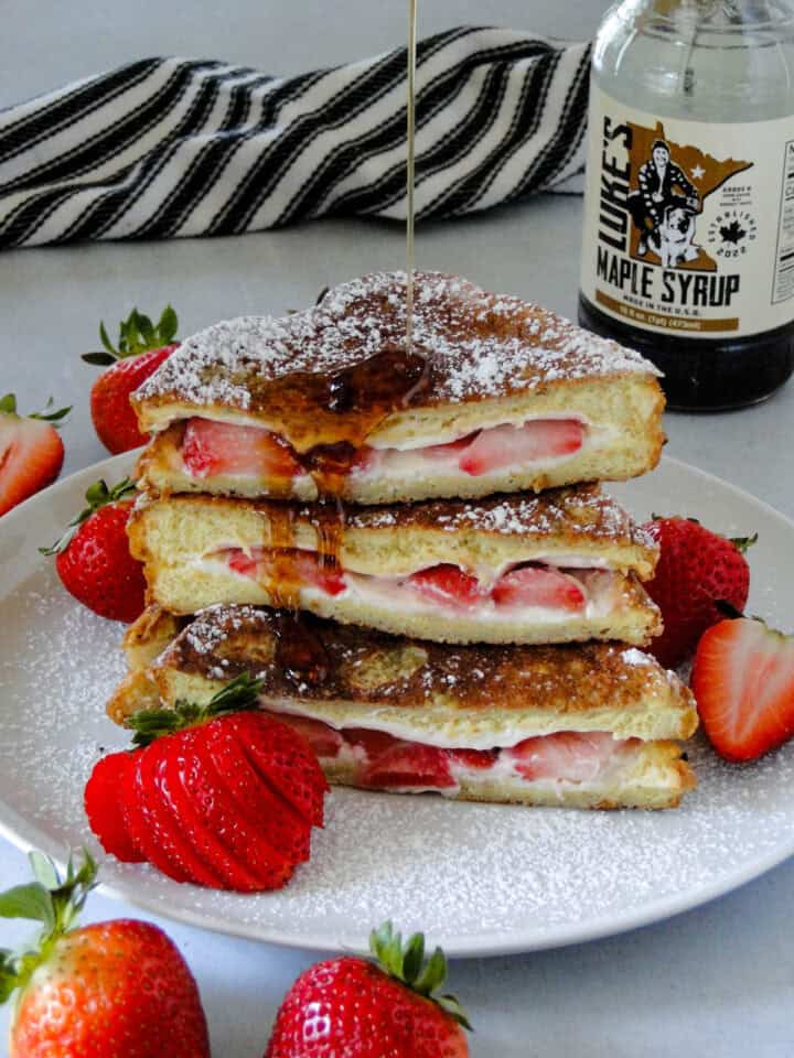 Side view of stuffed strawberry french toast cut in half, stacked and being drizzled with maple syrup on white round plate.