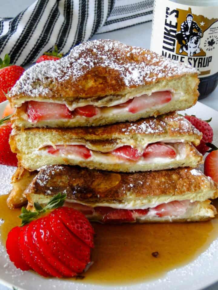 Close up side view of strawberries and cream stuffed french toast cut in half, stacked and drenched in maple syrup.