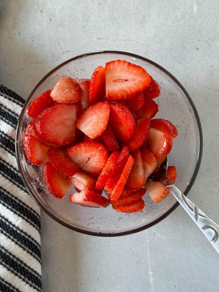 Sliced strawberries and sugar combined in glass mixing bowl.