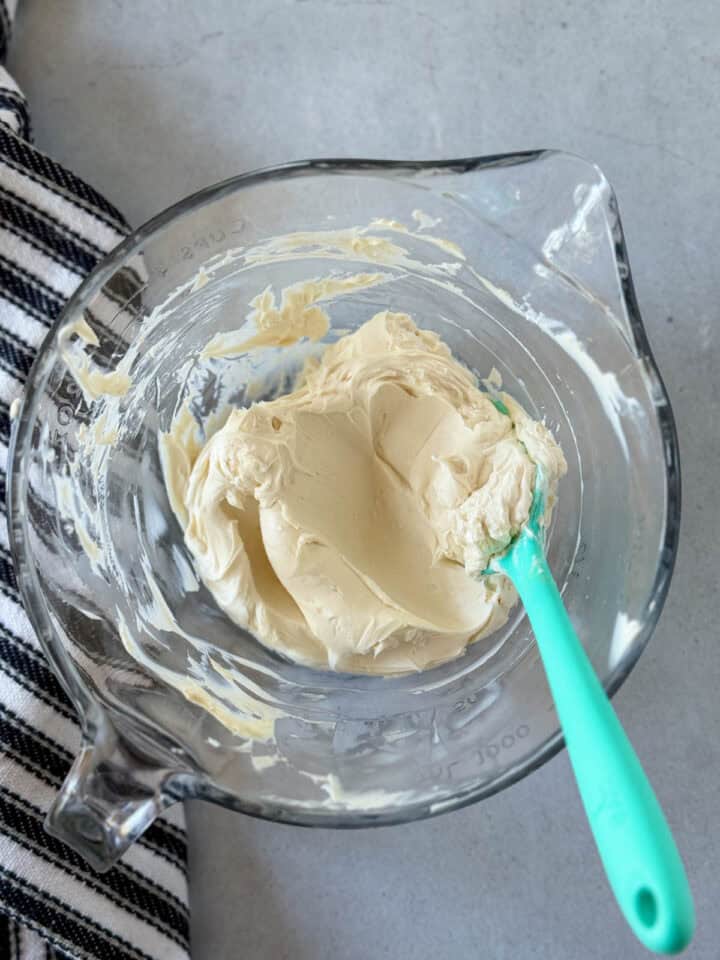 Cream cheese and vanilla extract mixed together in glass batter bowl with teal spatula.