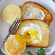 Egg in a hole on a plate with fork and the middle of the egg popped with yolk oozing out.