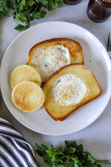 Top view of egg in a hole on white round plate with center pieces of bread next to them.
