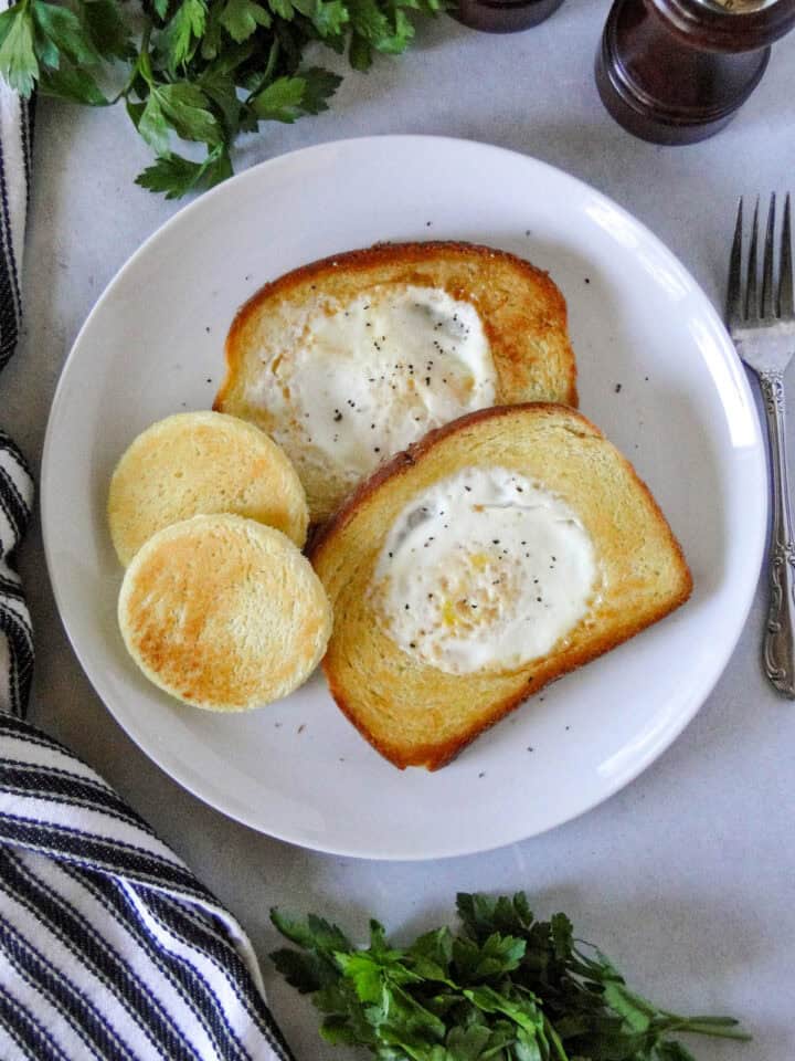Top view of egg in a hole on white round plate with center pieces of bread next to them.