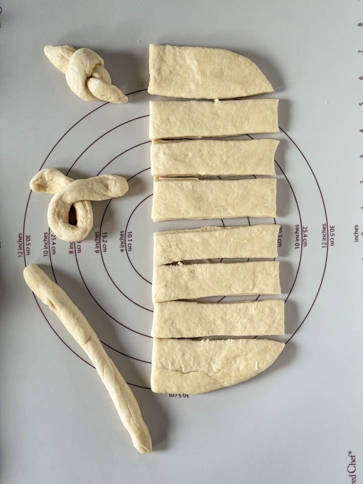 Bread dough cut into strips with one strip rolled into rope, another with rope end crossed over the other end and lastly tied into a knot all on a pastry mat.