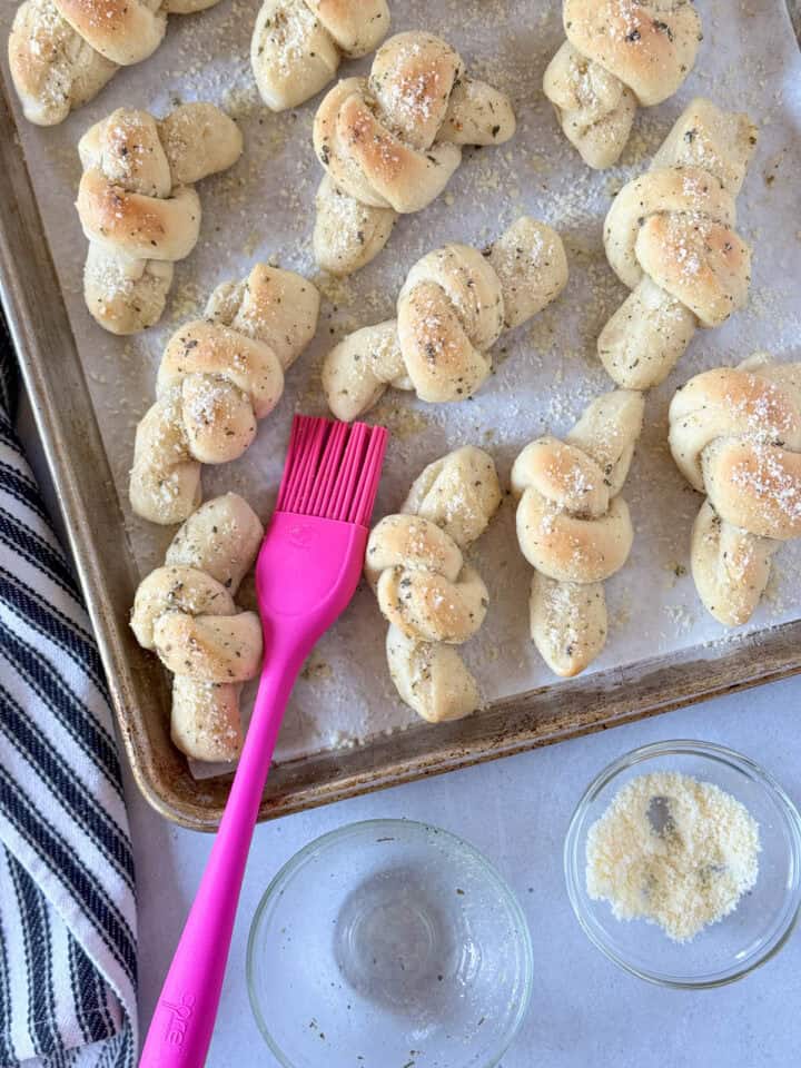 Baked garlic knots on sheet pan with pastry brush and bowl of garlic butter on the side.