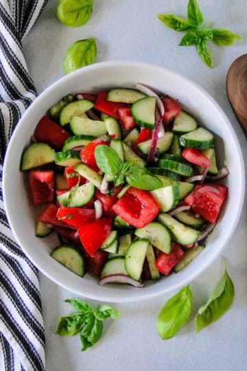 Top view of tomato cucumber basil salad in white serving bowl with wooden spoon to the right.